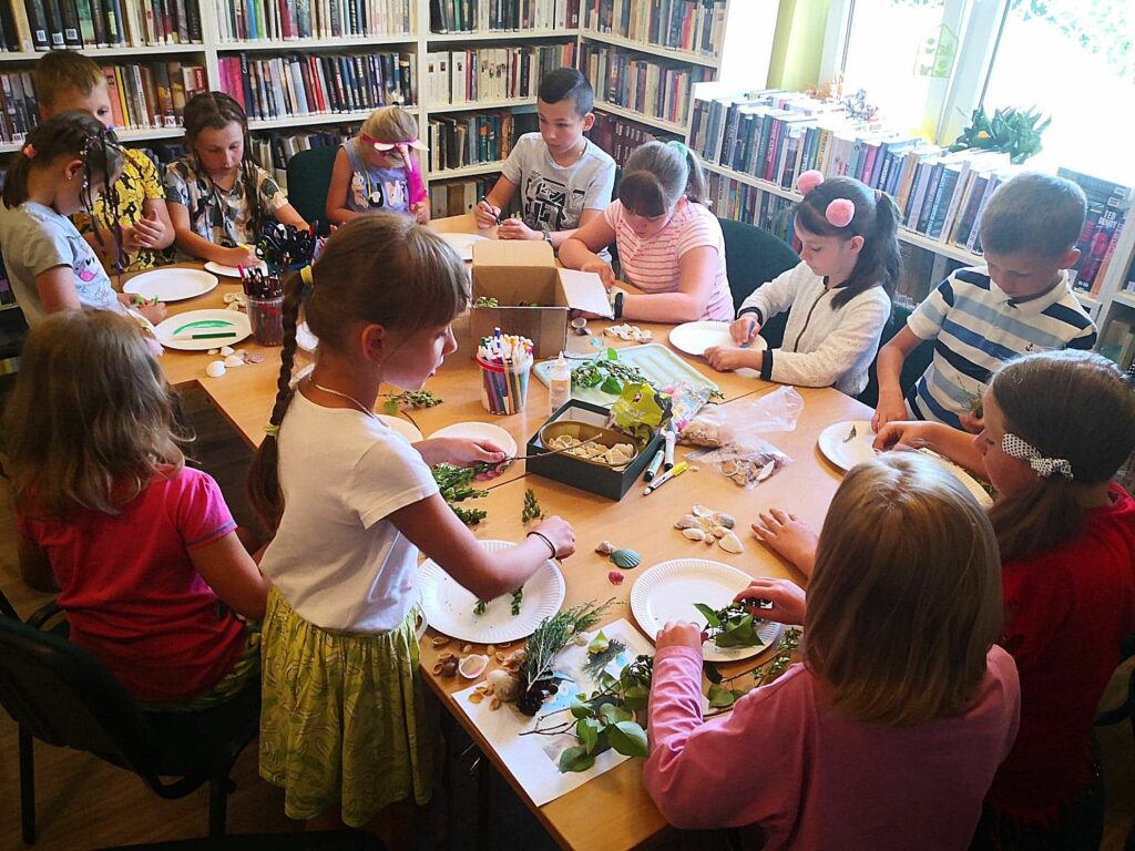 na zdjęciu widać dzieci siedzące przy stole w sali bibliotecznej wykonujące prace plastyczną