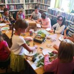na zdjęciu widać dzieci siedzące przy stole w sali bibliotecznej wykonujące prace plastyczną