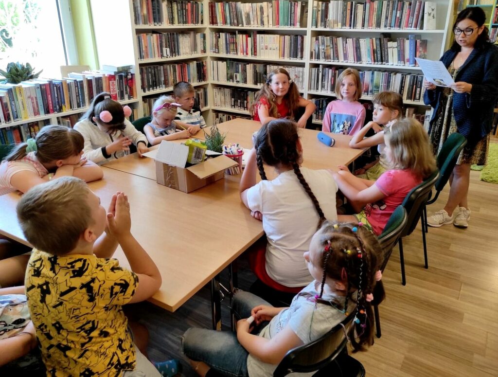 na zdjęciu widać dzieci siedzące przy stole i słuchające czytającej bibliotekarki, w tle widoczne są półki z książkami