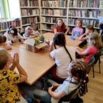 na zdjęciu widać dzieci siedzące przy stole i słuchające czytającej bibliotekarki, w tle widoczne są półki z książkami