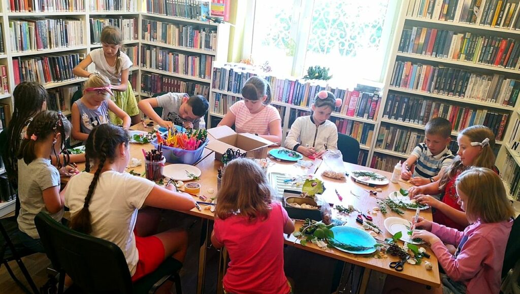 na zdjęciu widać dzieci siedzące wokół stołu i wykonujące prace plastyczną, widoczne jest również okno.