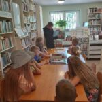 Grupa biorących udział w zajęciach dzieci siedzi przy stolikach i słucha czytanej im przez pana bibliotekarza książki pt. „Zawodowcy w świecie zwierząt”.