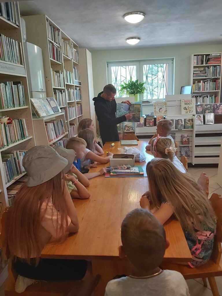 Grupa biorących udział w zajęciach dzieci siedzi przy stolikach i słucha czytanej im przez pana bibliotekarza książki pt. „Zawodowcy w świecie zwierząt”.