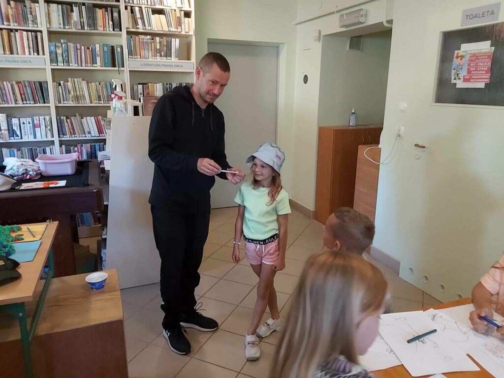 Na środku bibliotecznej sali stoi mała uśmiechnięta dziewczynka, a obok niej pan bibliotekarz, który pokazuje jej wylosowaną kartkę na której znajduje się owad, lub zwierzątko do naśladowania.