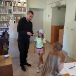 Na środku bibliotecznej sali stoi mała uśmiechnięta dziewczynka, a obok niej pan bibliotekarz, który pokazuje jej wylosowaną kartkę na której znajduje się owad, lub zwierzątko do naśladowania.