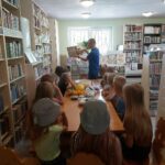 Grupa biorących udział w zajęciach dzieci siedzi przy stolikach i słucha czytanej im przez pana bibliotekarza książki pt. „Wanda szuka miłości”.