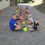 Na wyłożonym kostką placu obok biblioteki grupa dzieci i pan bibliotekarz siedzą w parach naprzeciwko siebie i grają w grę „Uszy, nos, koszulka”.