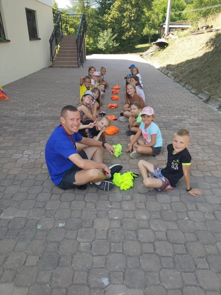 Na wyłożonym kostką placu obok biblioteki grupa dzieci i pan bibliotekarz siedzą w parach naprzeciwko siebie i grają w grę „Uszy, nos, koszulka”.