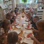 Wszystkie dzieci siedzą przy długim stole w bibliotece i ze smakiem jedzą czerwonego, soczystego arbuza.