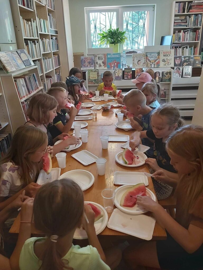 Wszystkie dzieci siedzą przy długim stole w bibliotece i ze smakiem jedzą czerwonego, soczystego arbuza.