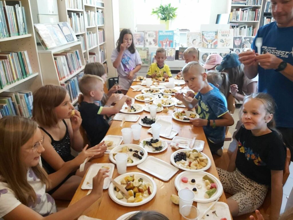 Owocowa uczta. Dzieci siedzą przy stole, każde z nich ma przed sobą talerzyk na którym znajduje się przygotowana przed momentem pyszna owocowa sałatka.