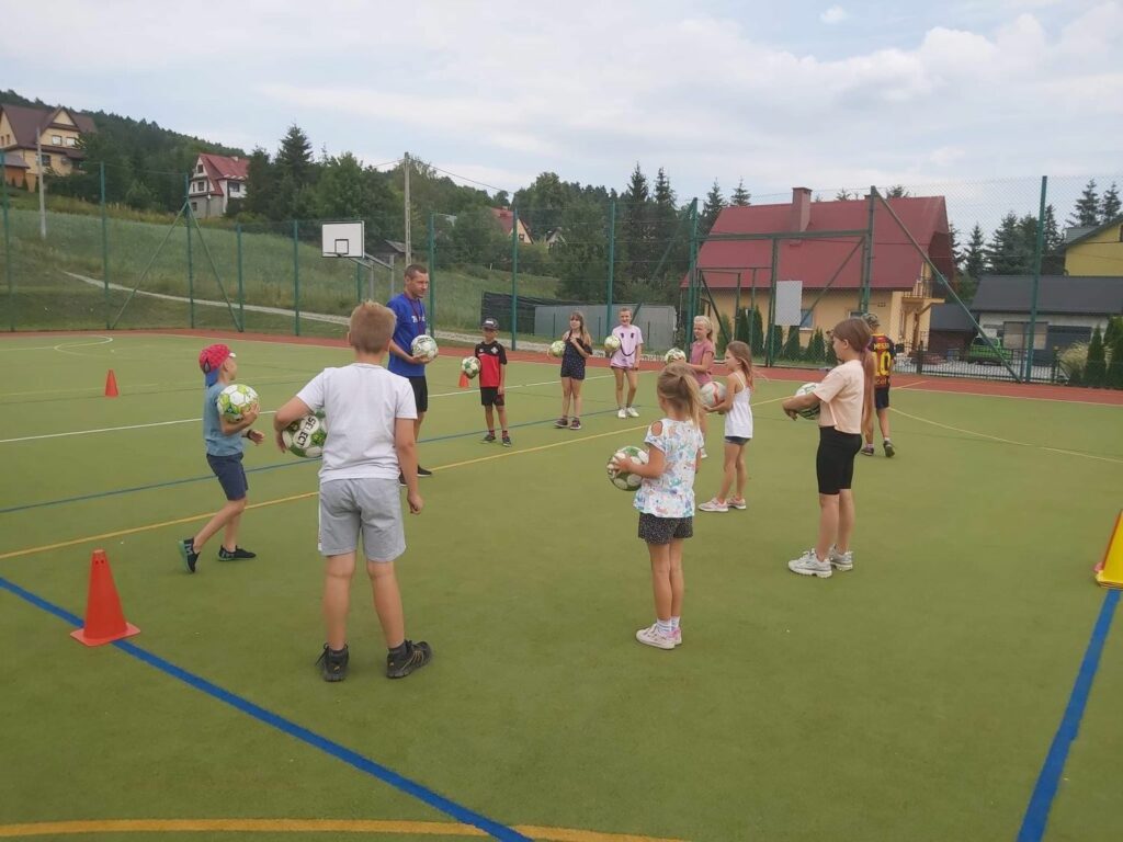 Dzieci znajdują się na boisku typu „Orlik”. Stoją w kole i każde trzyma w rękach piłkę. Na środku znajduje się pan bibliotekarz, który wyjaśnia im zasady zabawy „Polowanie na lisy”.
