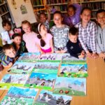Pd stołem stoją dzieci jesenaście uczestniczące w wakacjach z Biblioteką. Na stole rozłożone są prace wykonane przez dzieci.