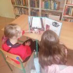 Dwie dziewczynki siedzą przy stole i czytają pełną kolorowych ilustracji książkę o przygodach dwóch sprytnych wiewiórek.