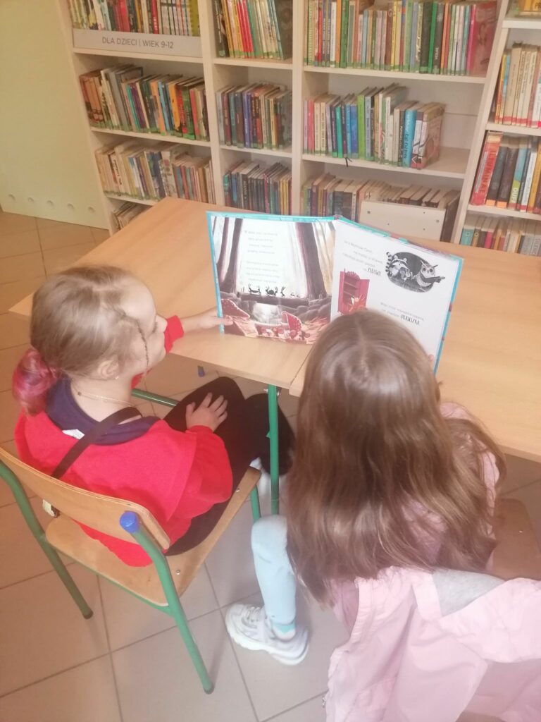 Dwie dziewczynki siedzą przy stole i czytają pełną kolorowych ilustracji książkę o przygodach dwóch sprytnych wiewiórek.