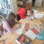 Dzieci siedzą przy bibliotecznym stole i kolorują na kartkach swoje wiewiórki. Za moment przy pomocy pomarańczowej bibuły i kleju zaczną ozdabiać ich ogonki.