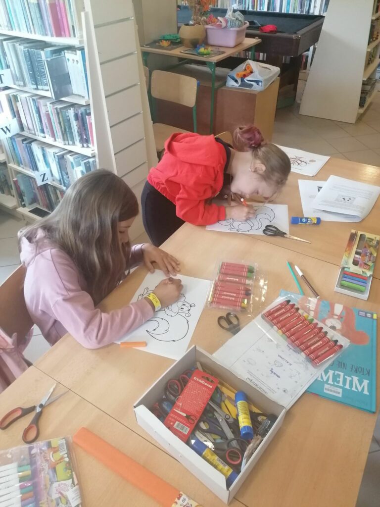 Dzieci siedzą przy bibliotecznym stole i kolorują na kartkach swoje wiewiórki. Za moment przy pomocy pomarańczowej bibuły i kleju zaczną ozdabiać ich ogonki.