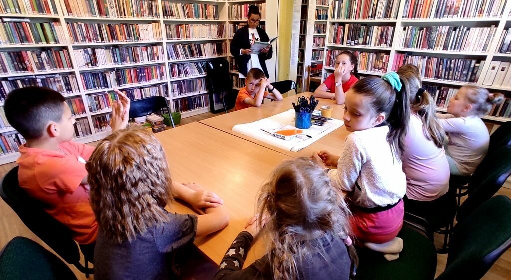 na zdjęciu widać sale biblioteczną oraz siedzące wokół stolika dzieci, które słuchają czytanej im przez bibliotekarkę książki.
