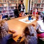 na zdjęciu widać sale biblioteczną oraz siedzące wokół stolika dzieci, które słuchają czytanej im przez bibliotekarkę książki.