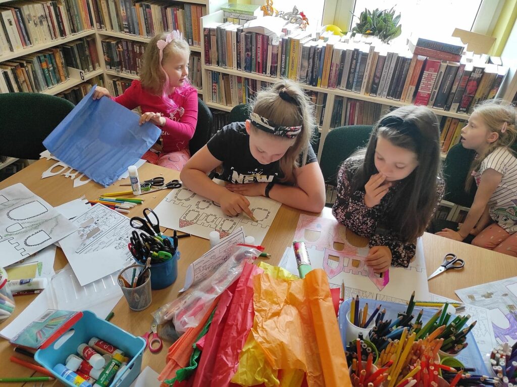 na zdjęciu widać sale biblioteczną, regały z książkami, na środku sali stoi stół, przy stole siedzą dzieci, wykonują pracę plastyczną