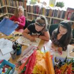 na zdjęciu widać sale biblioteczną, regały z książkami, na środku sali stoi stół, przy stole siedzą dzieci, wykonują pracę plastyczną