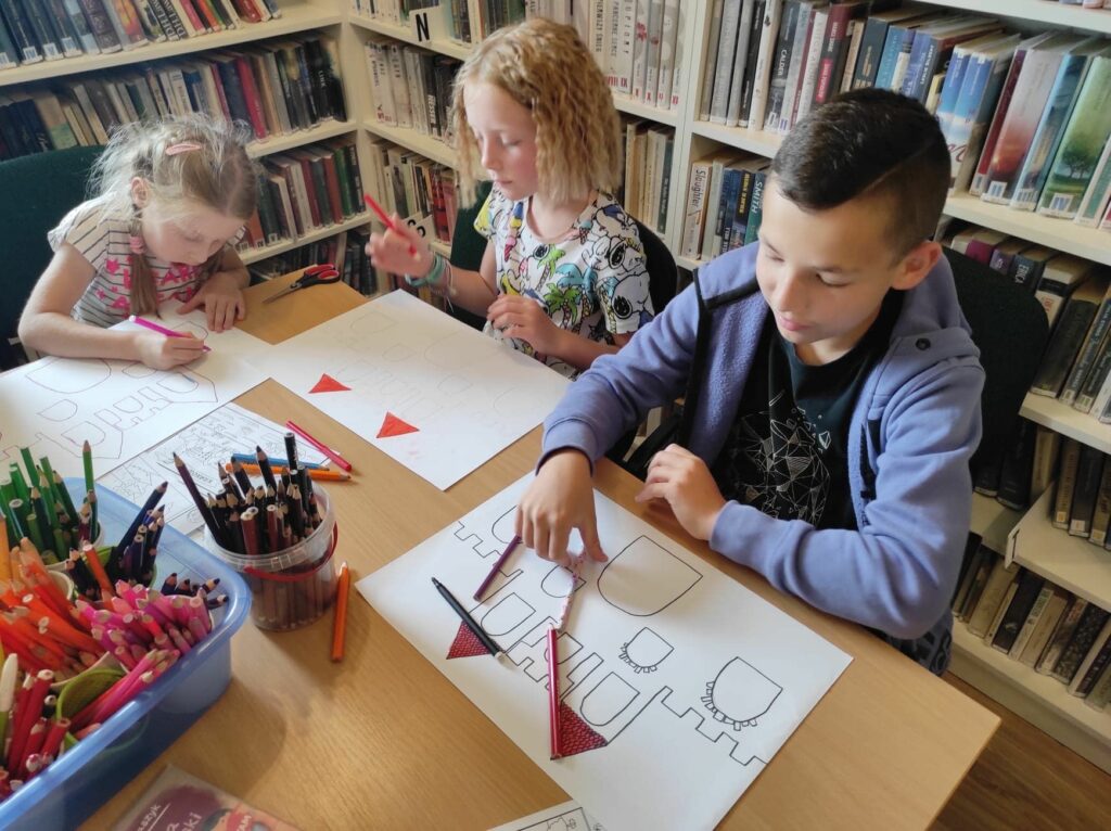 na zdjęciu widać sale biblioteczną, regały z książkami, na środku sali stoi stół, przy stole siedzą dzieci, wykonują pracę plastyczną