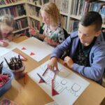 na zdjęciu widać sale biblioteczną, regały z książkami, na środku sali stoi stół, przy stole siedzą dzieci, wykonują pracę plastyczną