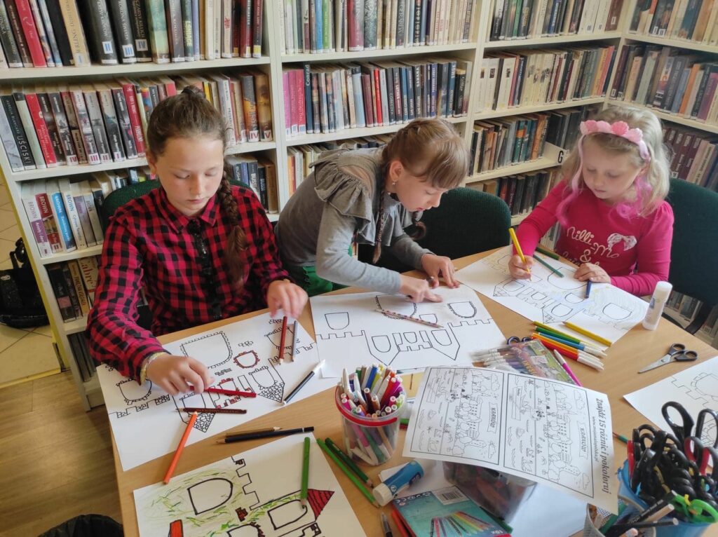 na zdjęciu widać sale biblioteczną, regały z książkami, na środku sali stoi stół, przy stole siedzą dzieci, wykonują pracę plastyczną