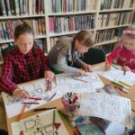 na zdjęciu widać sale biblioteczną, regały z książkami, na środku sali stoi stół, przy stole siedzą dzieci, wykonują pracę plastyczną