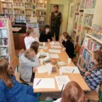Grupa dzieci siedzi przy stolikach w bibliotecznej sali i słucha ubranego na zielono Leśnika opowiadającego im o swojej pracy.