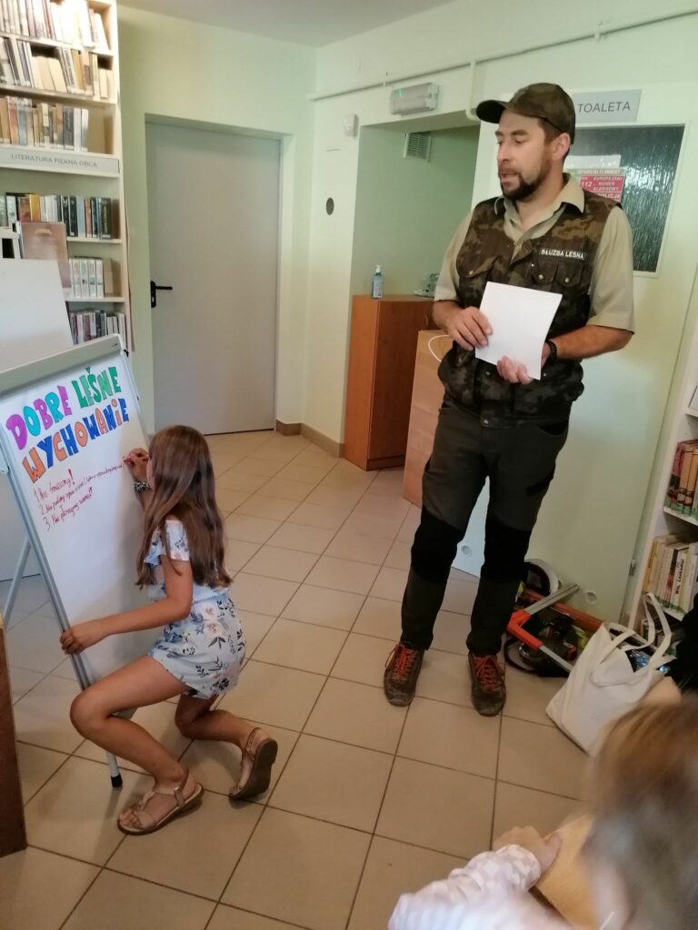 Jedna z dziewczynek pisze na wiszącym na stelażu plakacie, czego nie można robić w lesie. Obok niej stoi pan Leśnik i opowiada m.in. o zakazie śmiecenia w lasach.