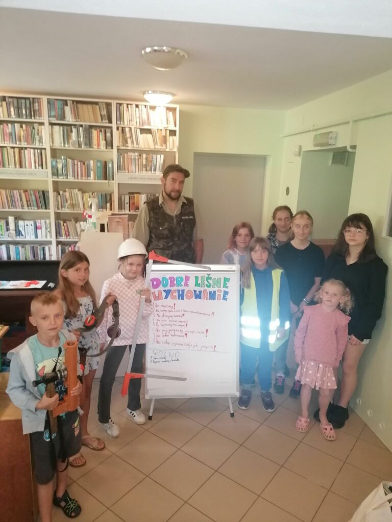 Wszystkie dzieci ustawiły się wokół pana Leśnika. Pomiędzy nimi stoi narysowany wspólnie wielki plakat pt. „Dobre leśne wychowanie”. Uczestnicy trzymają w rękach podstawowy sprzęt używany w pracy Leśnika czyli np. kask ochronny, kulpę do pomiarów i numerator - przyrząd do oznaczania ściętych drzew.