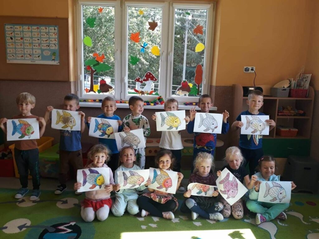 Grupa przedszkolaków ustawiła się do zdjęcia. Każde dziecko trzyma w rękach pięknie pokolorowany rysunek „Złotej Rybki”.