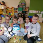 Piątka dzieci siedzi na wykładzinie na ziemi. Chłopiec w środku trzyma grę Duszki. Z tyłu za nimi kolorowe meble przedszkolne oraz zabawki.