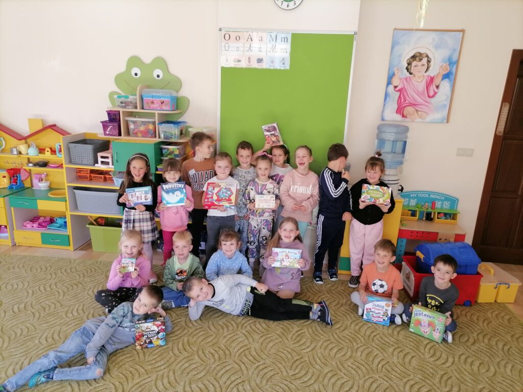 W sali w przedszkolu stoją dzieci ustawione do zdjęcia. Dziesiatka dzieci trzyma w ręku gry planszowe. Dziesiątka dzieci stoi natomiast ósemka siedzi na ziemi.