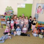 W sali w przedszkolu stoją dzieci ustawione do zdjęcia. Dziesiatka dzieci trzyma w ręku gry planszowe. Dziesiątka dzieci stoi natomiast ósemka siedzi na ziemi.