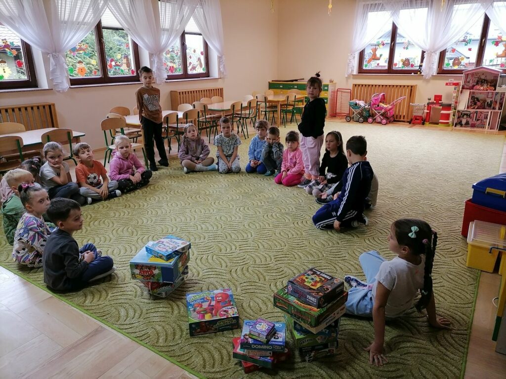 W sali w przedszkolu grupa dzieci siedzi w kółeczku. jedna dziewczynka i jeden chłopiec stoi. Wokół zabawki kolorowe. Obok dzieci na ziemi kilkanaście pudełek z grami planszowymi.