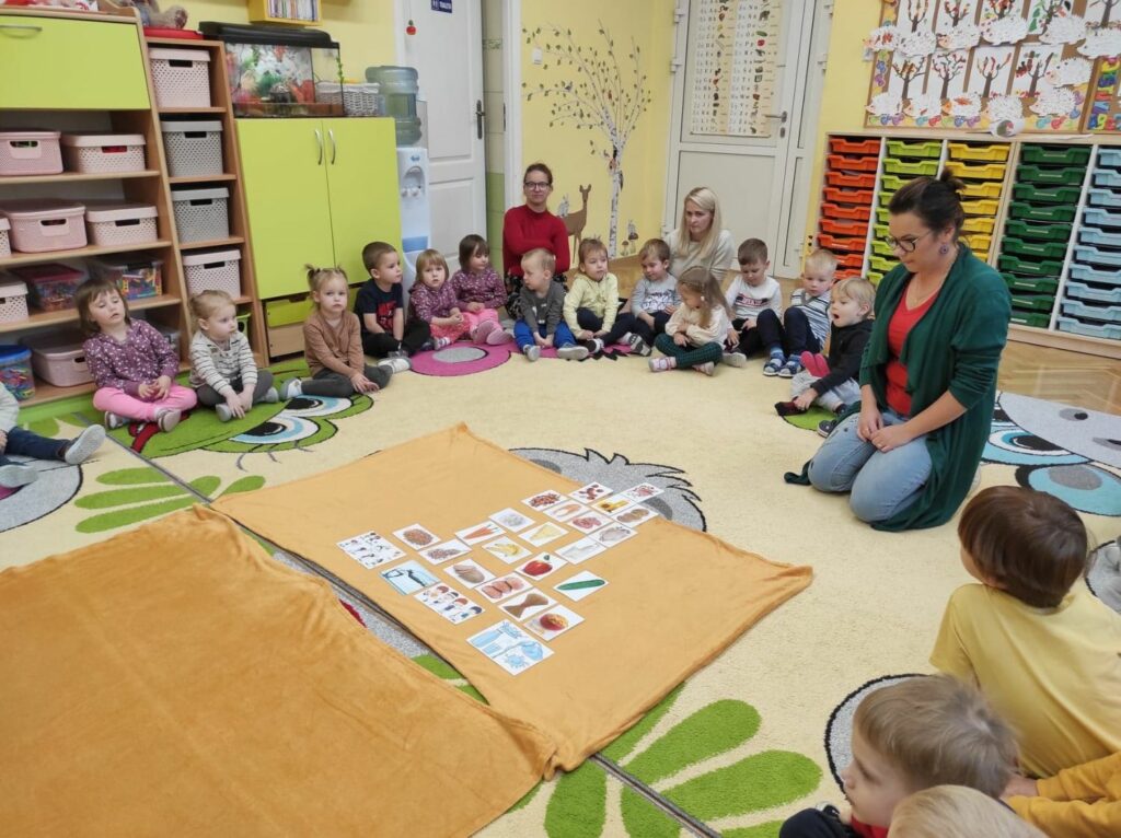 na zdjęciu widać kolorową sale przedszkolną , dzieci siedzące na dywanie oraz stojącą i czytającą bibliotekarkę