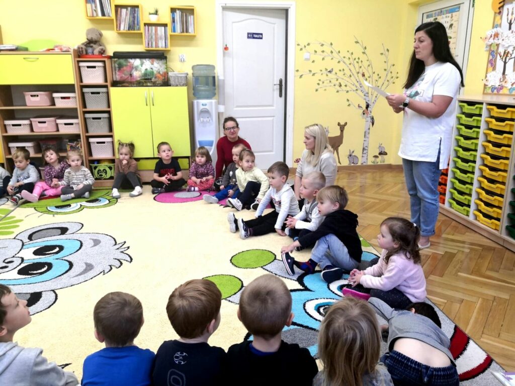 na zdjęciu widać przedszkolanki oraz dzieci siedzące na kolorowym dywanie, w tle widać kolorowe dekoracje przedszkolne.