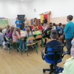 Pan bibliotekarz stoi przy powieszonej na ścianie wielkiej mapie Polski i czyta siedzącym w ławkach dzieciom legendy polskie.