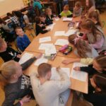 Grupa uśmiechniętych dzieci siedzi wokół złączonych razem ławkach i rozwiązuje na kartkach podchwytliwe „wykreślanki”.