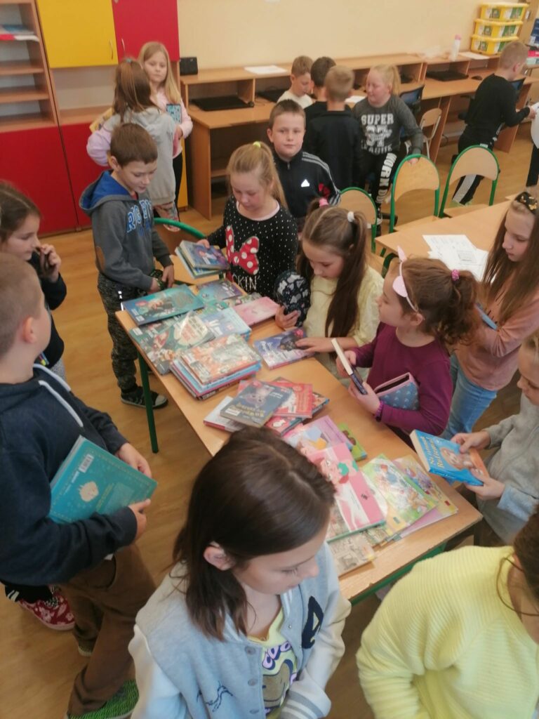 Uczniowie jednej z biorących udział w spotkaniu klasy stoją tłumnie wokół stolika na którym znajduje się mnóstwo kolorowych książek. Dzieci wybierają lektury, które za chwilę wypożyczą.