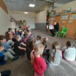 Spora grupa przedszkolaków siedzi wygodnie na dywanie pod ścianą i słucha siedzącego przed nimi na krześle pana bibliotekarza, który czyta im kolorową bajkę.