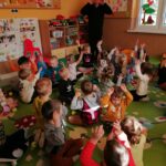 Grupa dzieci siedzi wygodnie na dywanie i wykonuje demonstrowane im przez pana bibliotekarza ćwiczenia.