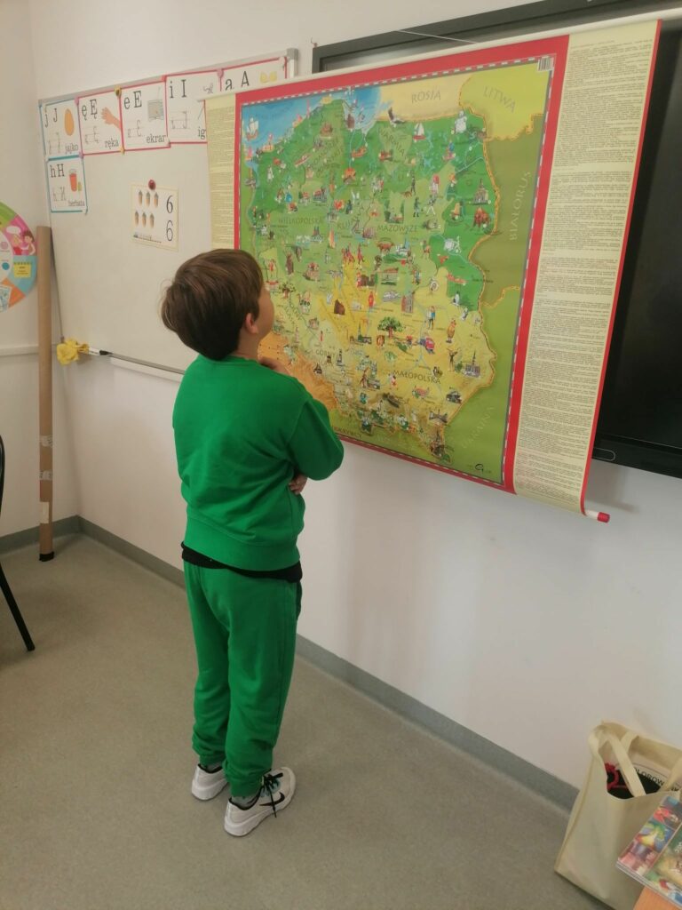 Na ścianie obok tablicy wisi wielka kolorowa mapa naszego kraju. Tuż przy niej stoi ubrany w zielony dres chłopiec i skoncentrowany szuka najgłębszego polskiego jeziora Hańcza.