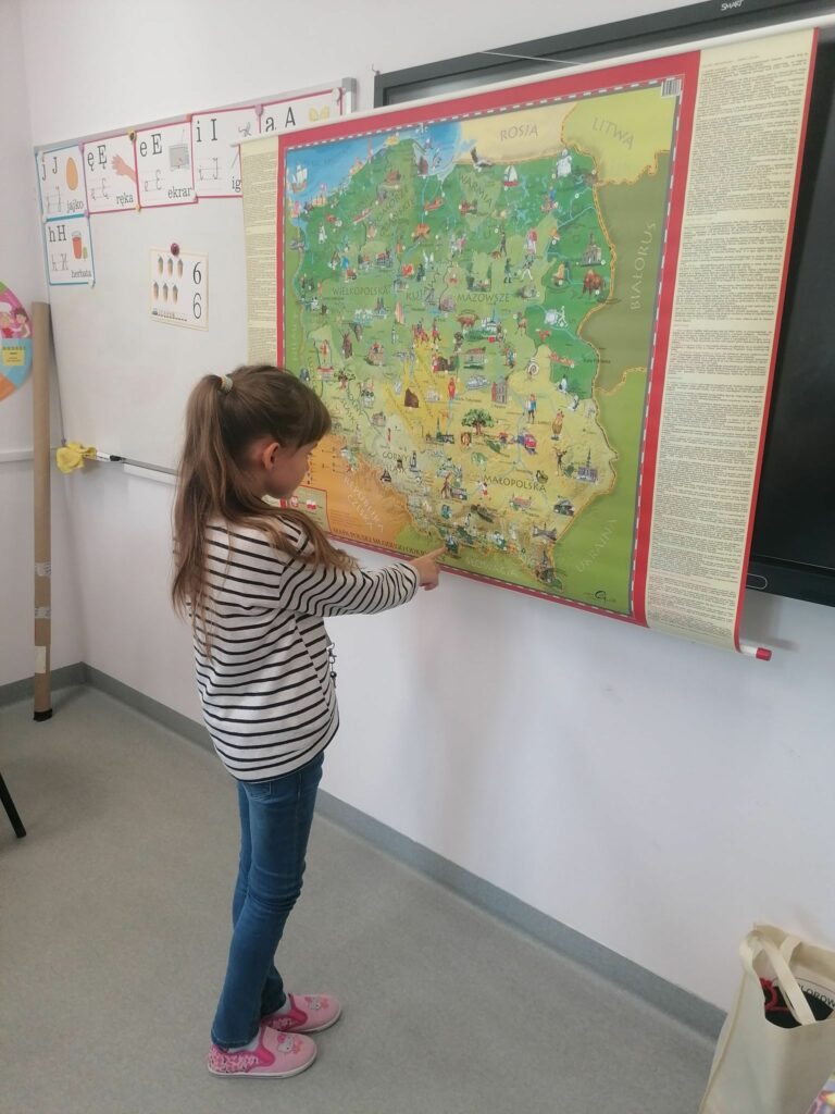 Przy wielkiej kolorowej mapie Polski stoi dziewczynka i pewnie wskazuje palcem najwyższy szczyt naszego kraju Rysy.