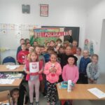 Wszystkie dzieci biorące udział w zajęciach ustawiły się pod tablicą do pamiątkowego zdjęcia. Stojący z tyłu dwaj uśmiechnięci chłopcy trzymają nad głowami biało- czerwony szalik z napisem „Polska”.