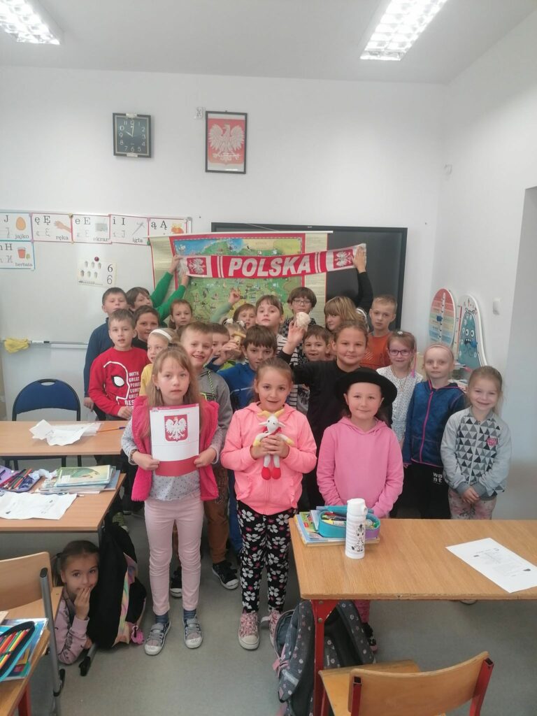 Wszystkie dzieci biorące udział w zajęciach ustawiły się pod tablicą do pamiątkowego zdjęcia. Stojący z tyłu dwaj uśmiechnięci chłopcy trzymają nad głowami biało- czerwony szalik z napisem „Polska”.