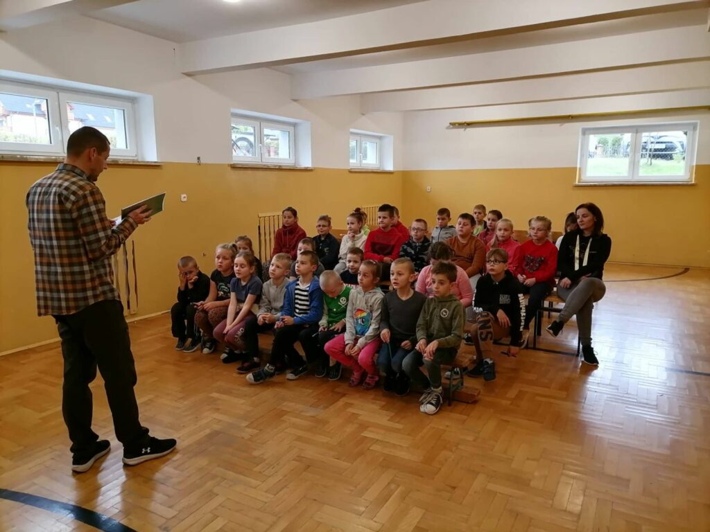 Wszystkie dzieci z klas I, II, III i IV zebrały się na szkolnej sali gimnastycznej i słuchają z zainteresowaniem książki pt. „Nasza paczka i niepodległość”.