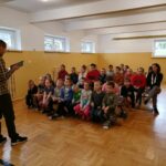 Wszystkie dzieci z klas I, II, III i IV zebrały się na szkolnej sali gimnastycznej i słuchają z zainteresowaniem książki pt. „Nasza paczka i niepodległość”.